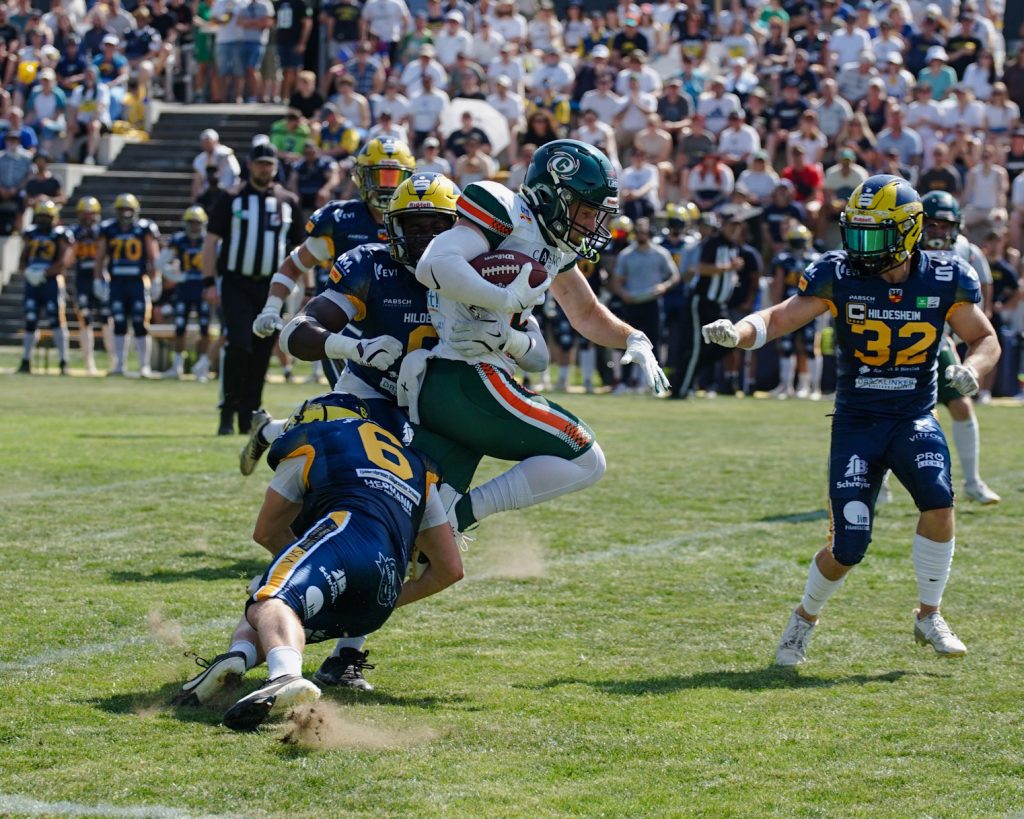 Hurricanes Receiver Chandler Smith (im weißen Trikot und grüner Hose) trägt den Football im rechten Arm und zwei Hildesheimer in ihren blauen Uniformen versuchen ihn zu stoppen. Einer unten an den Beiden und der andere hat einen Arm um Smiths Taille. Im Hintergrund sieht man die gut besetzte Tribüne auf dem Hildesheimer Homefield.