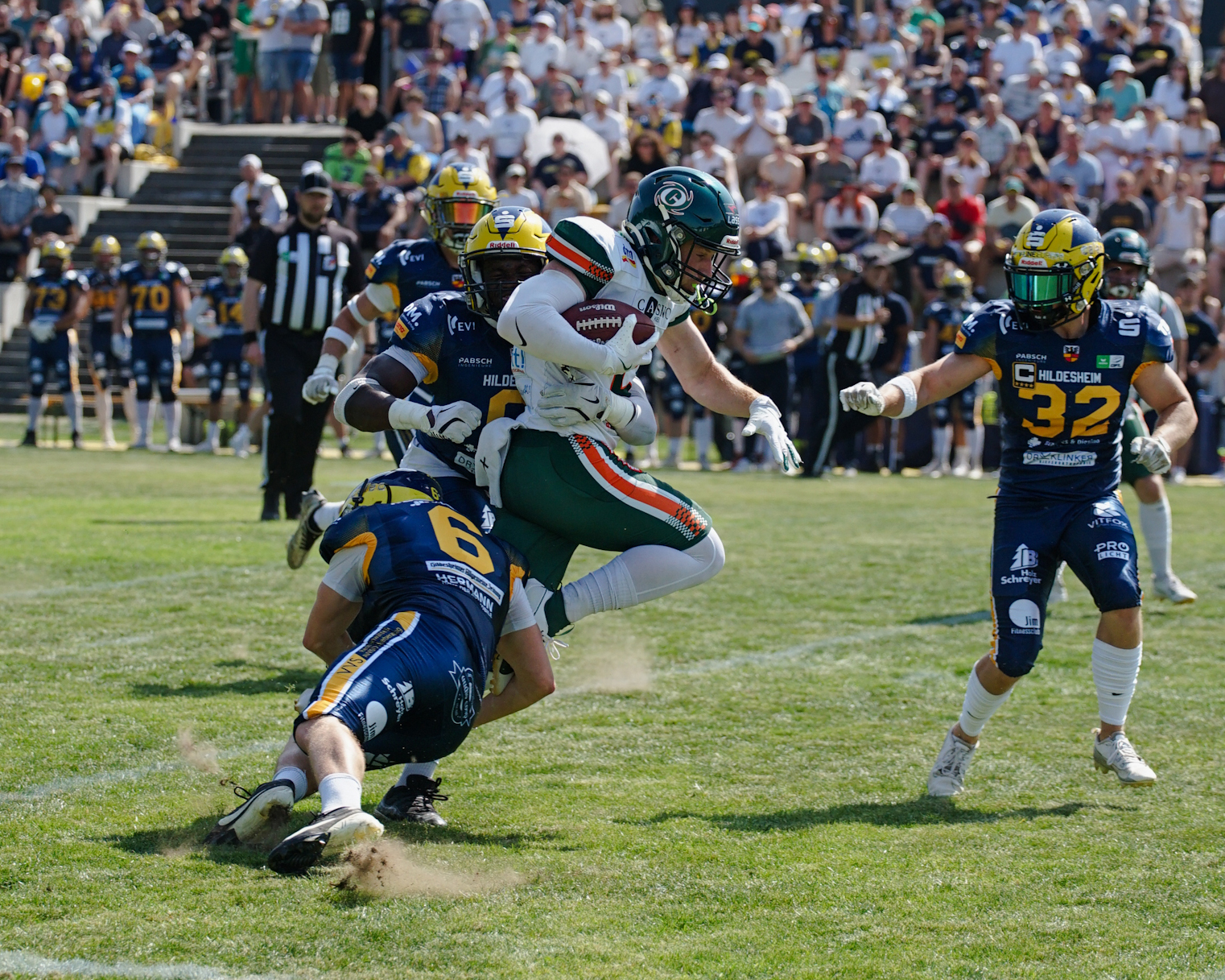 Hurricanes Receiver Chandler Smith (im weißen Trikot und grüner Hose) trägt den Football im rechten Arm und zwei Hildesheimer in ihren blauen Uniformen versuchen ihn zu stoppen. Einer unten an den Beiden und der andere hat einen Arm um Smiths Taille. Im Hintergrund sieht man die gut besetzte Tribüne auf dem Hildesheimer Homefield.